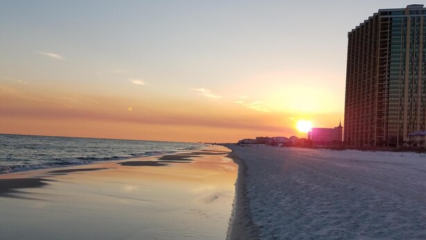 A beach with a sunset in the background, a sandy shore, and a tall building to the right. - LOFT UNIT ON THE BEACH (Orange Beach)
