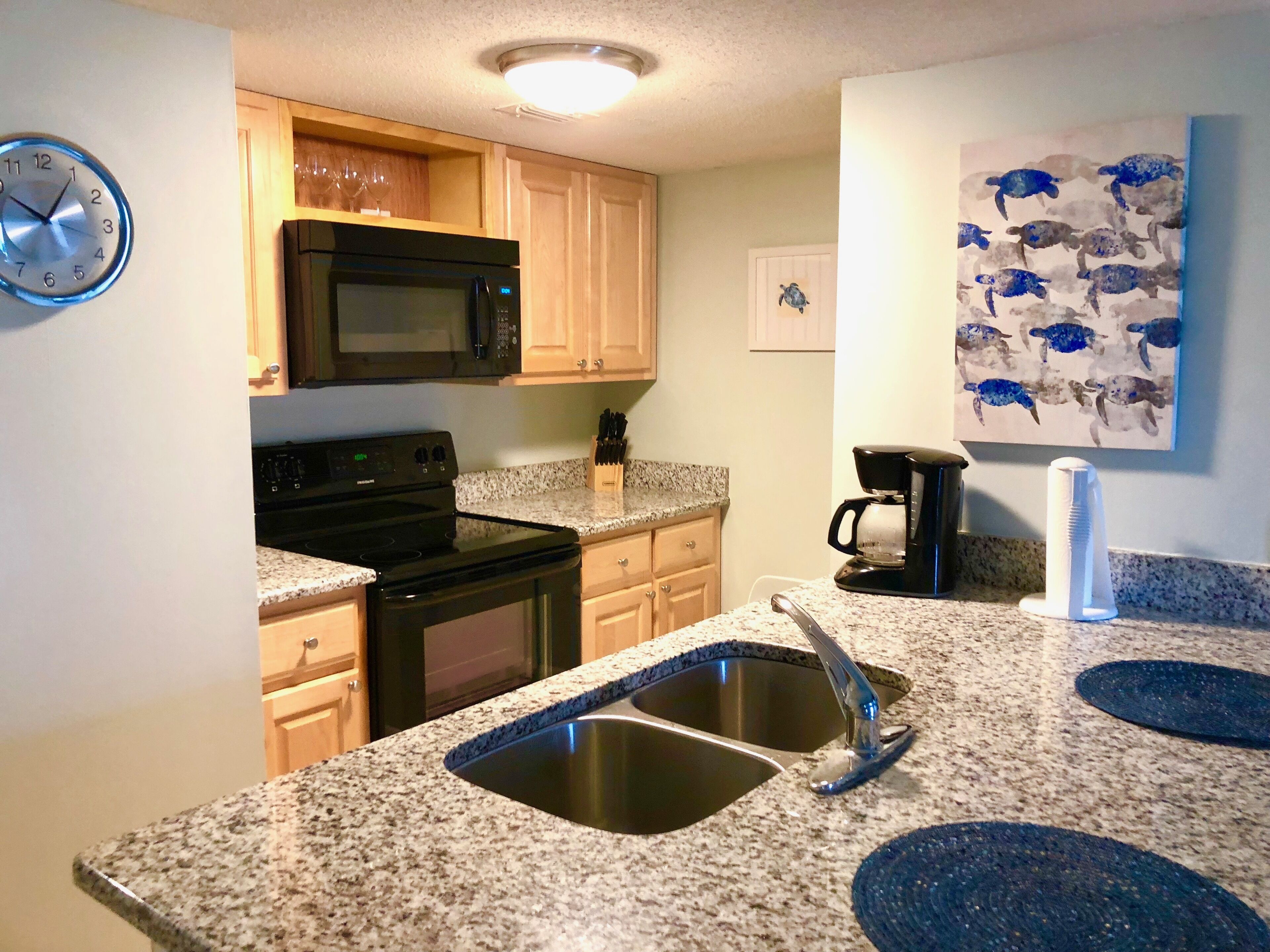 A kitchen with granite countertops, stainless steel appliances, wooden cabinets, a microwave, a coffee maker, and a wall clock.