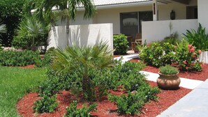 A house with a covered porch, a well-maintained garden with various shrubs and a palm tree, and a red mulch pathway. - Quick Walk To Beach, Restaurants, Shops, Movies. Updated, Spacious. Private Pool (Marco Island)