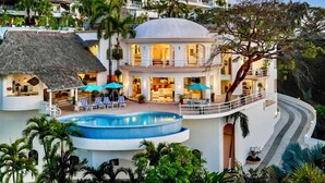 A multi-story resort with a swimming pool, lounge area, and thatched-roof structure.