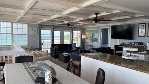 Een ruime woonkamer met een eettafel, banken, een keuken met een televisie en grote ramen met uitzicht op het strand.