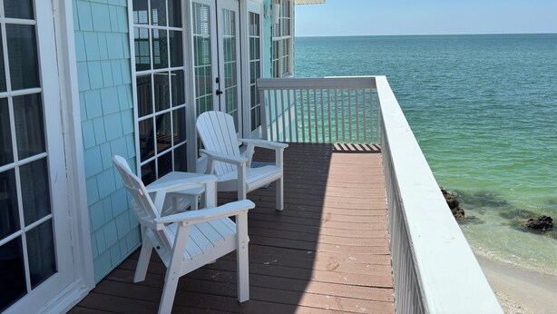 Een houten terras met twee witte Adirondack-stoelen, een glazen deur en uitzicht op de oceaan.