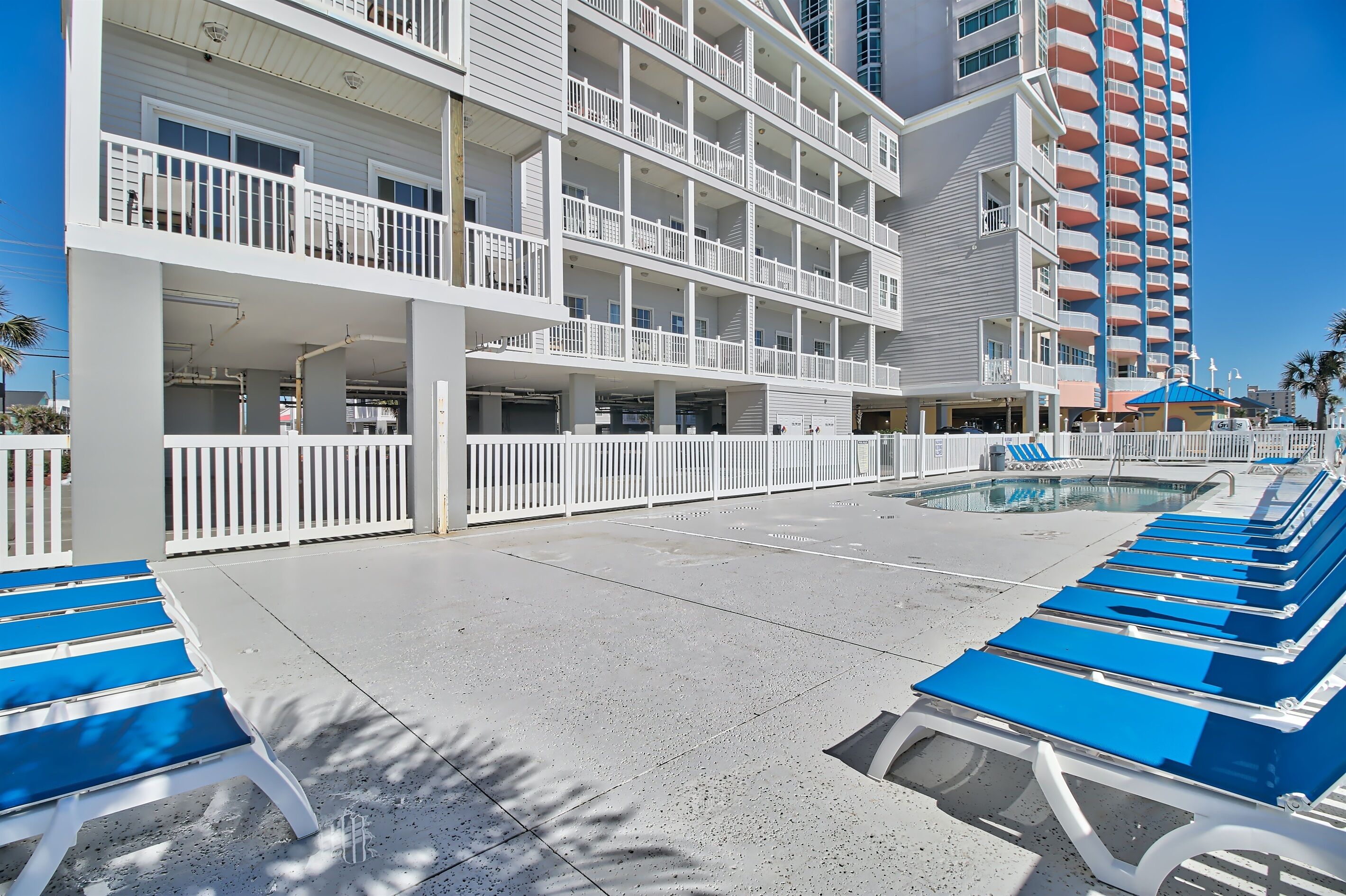 A hotel with multiple balconies, a pool area with lounge chairs, and a clear blue sky.