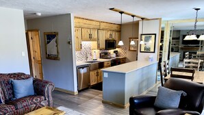 A modern kitchen with wooden cabinets, a central island, and pendant lighting. There is a dining area with wooden tables and chairs.