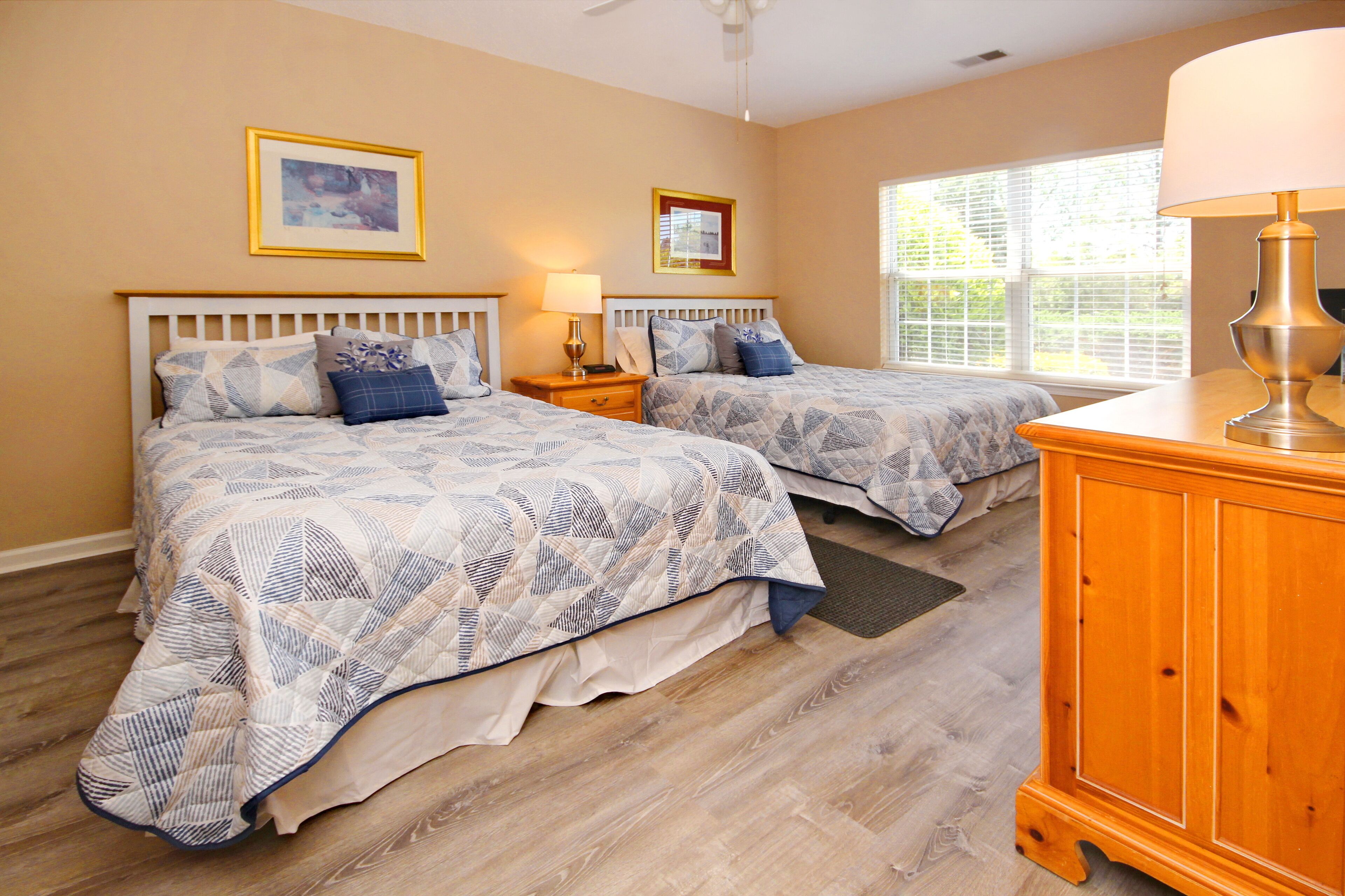 A bedroom with two beds, wooden headboards, a nightstand with a lamp, a wooden dresser, and a window with blinds.