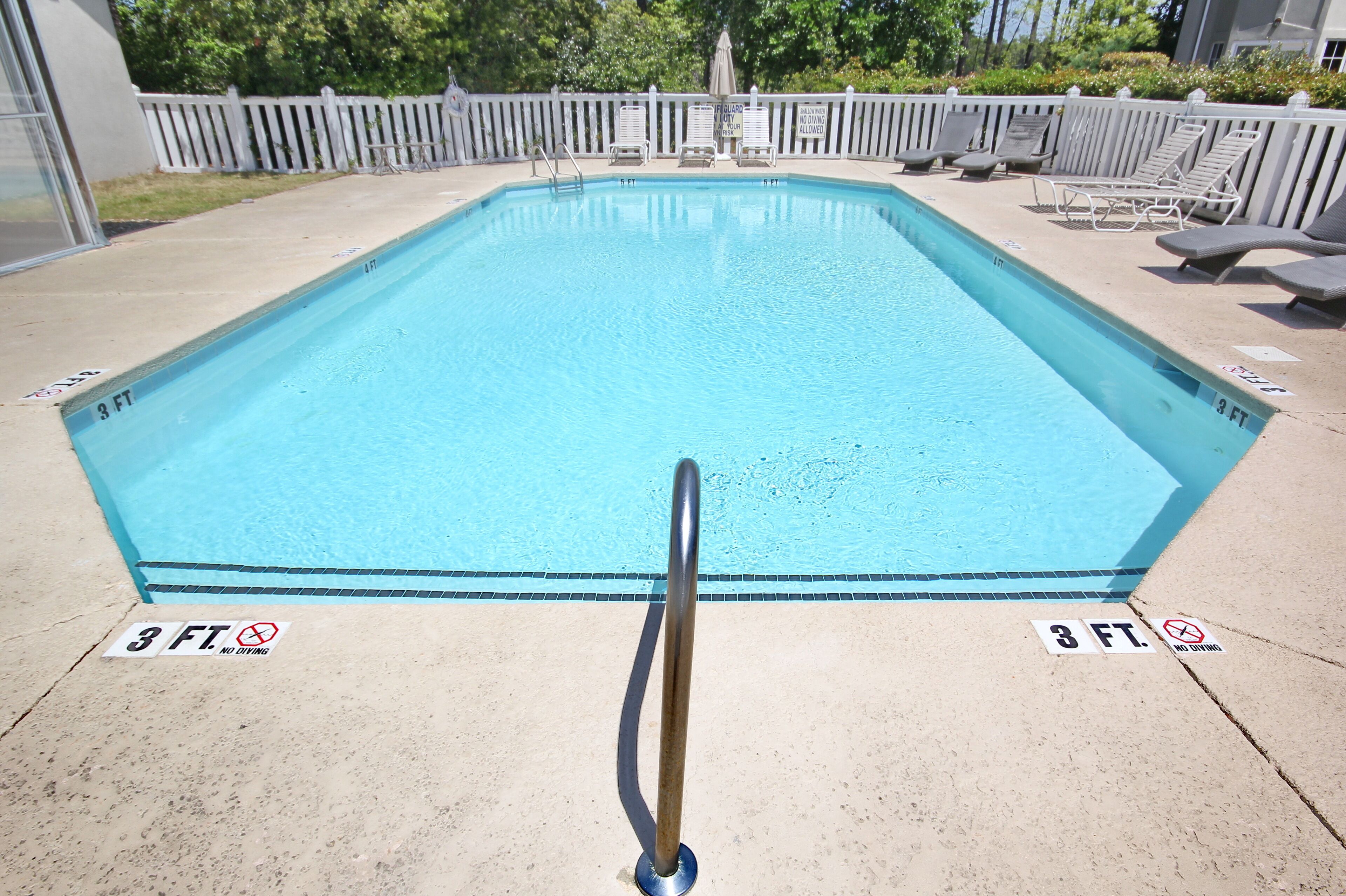 Une piscine rectangulaire avec une rambarde de sécurité en métal au premier plan. Des chaises longues et des parasols sont disposés le long du bord de la piscine.