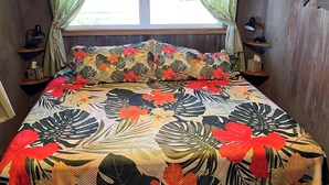 Una cama con cubrecama floral, una ventana con cortinas, una lámpara de noche y una mesita con lámpara y caja de pañuelos.