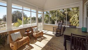 A sunroom with wicker furniture, a dining table, and large windows offering a view of palm trees and a building.