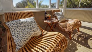 A wicker lounge chair with a patterned pillow, a small table with a bottle and glasses, and a view of palm trees through large windows.