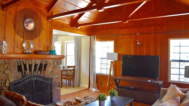 Interior acolhedor de uma cabana de toras, com lareira de pedra, televisão e área de jantar.