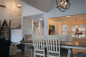 A dining area with a wooden table and white chairs, a staircase, and a kitchen visible in the background. - One-of-a-Kind Luxury Lakefront Home; Waterfall Landscape; Exclusive Boat Rental (Lake Ozark)