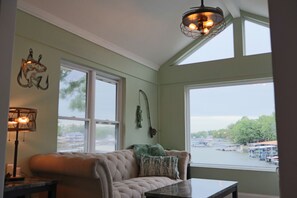 A room with a large window, a cushioned sofa, a table, and a lamp. - One-of-a-Kind Luxury Lakefront Home; Waterfall Landscape; Exclusive Boat Rental (Lake Ozark)