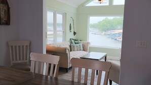 A room with a sofa, two chairs, a small table, a window with a view of boats, and a wall-mounted light fixture.