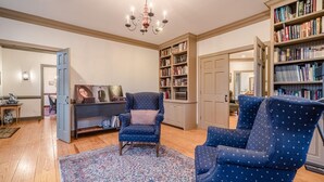 A living room with a blue armchair, a patterned rug, and a bookshelf filled with books. - Special Rates-Enjoy Hot Springs in Our Colonial Williamsburg Reproduction Home (Hot Springs)