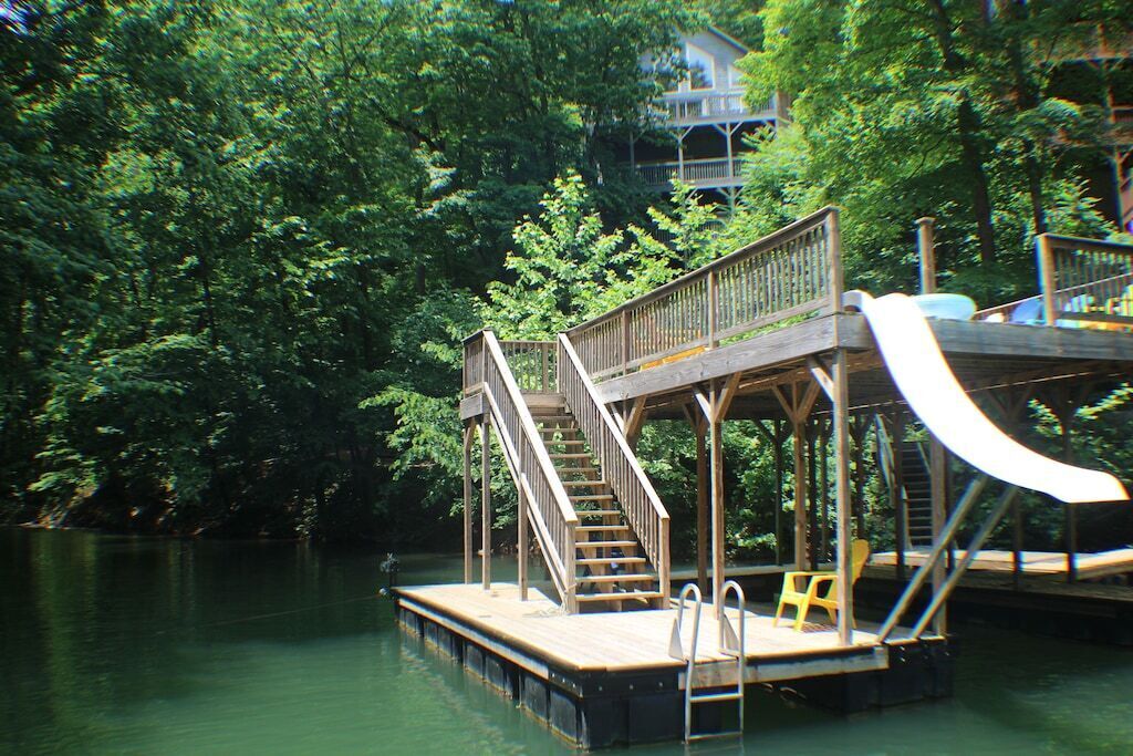 A wooden dock with a slide and stairs leading to a platform, surrounded by dense greenery.