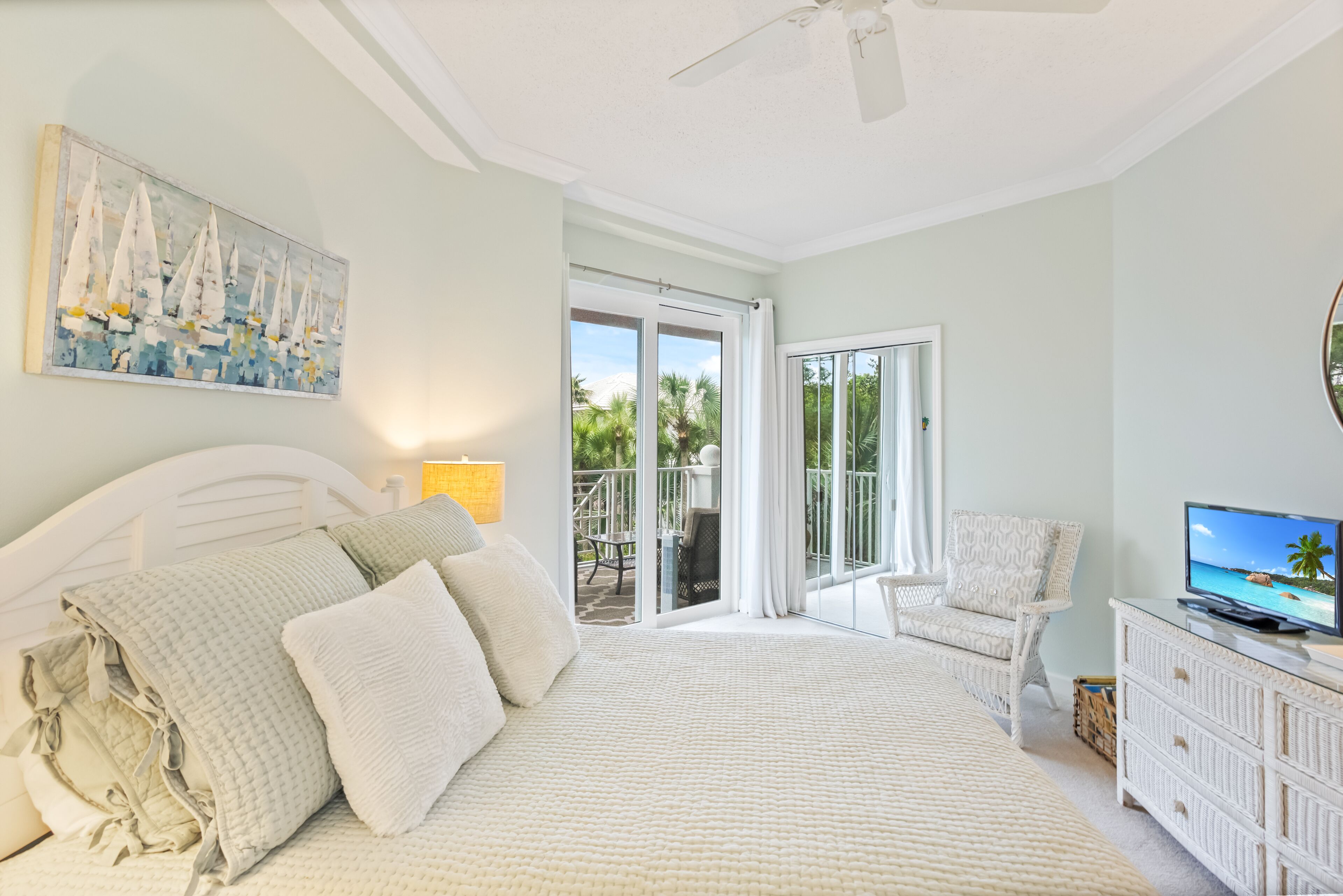 A bedroom with a bed, a chair, a television, and a balcony with a view of palm trees.