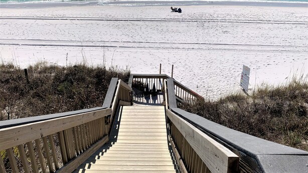 En træpromenade der fører til en strand med klart turkis vand og en sandbund.