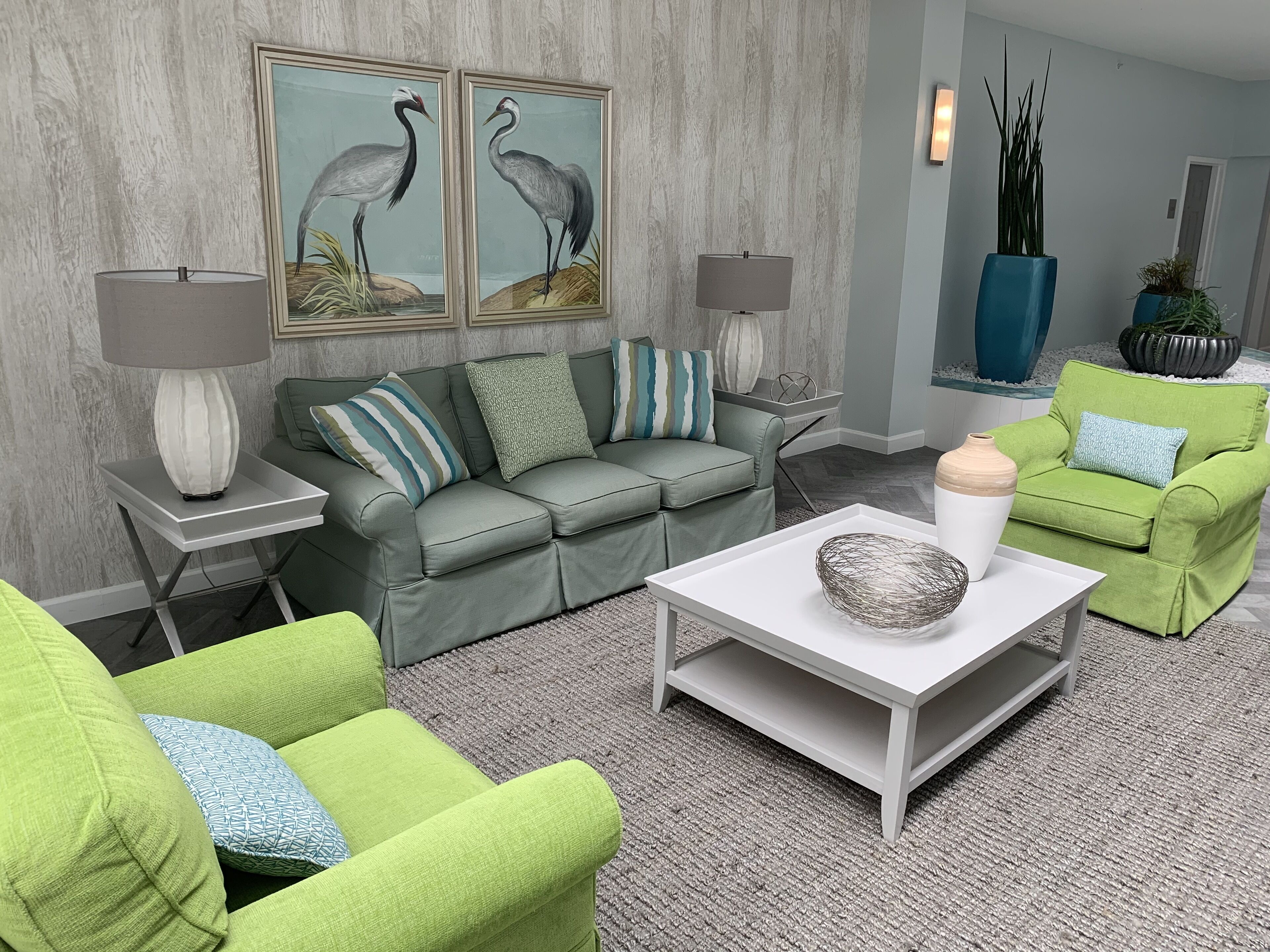 Una sala de estar moderna con un sofá gris, sillones verdes, una mesa de centro blanca y dos cuadros enmarcados de aves.