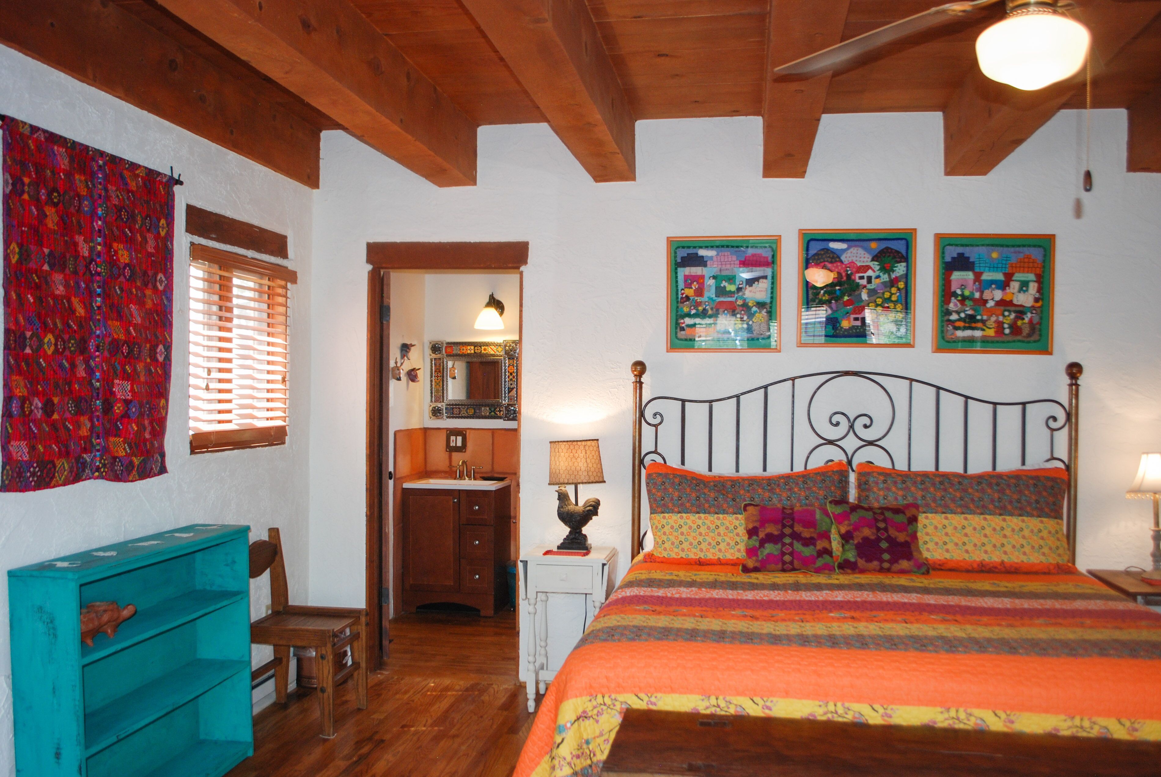 Une chambre à coucher avec un lit, des poutres en bois au plafond, une fenêtre avec des stores, une petite table avec une lampe, une chaise et une couverture colorée.
