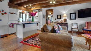 A spacious living room with a large brown sofa, a wooden ceiling, and a kitchen area with white cabinets and a blue countertop.