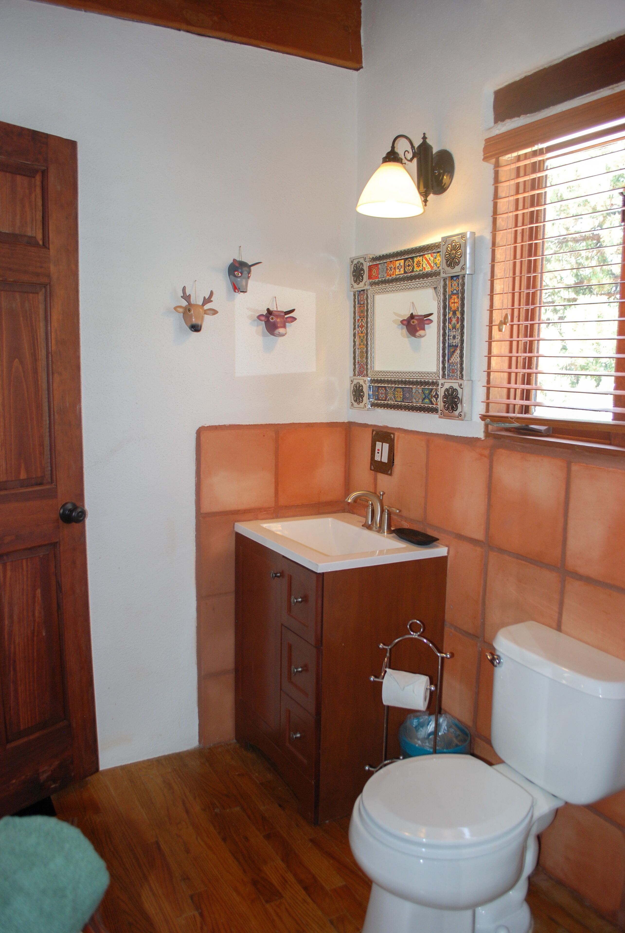 Un baño con puerta de madera, un lavamanos con espejo arriba, un inodoro y una ventana con persianas.