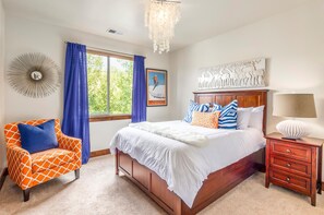 Une chambre à coucher avec un grand lit, une chaise, une table de chevet, un lustre, une fenêtre avec des rideaux et un élément décoratif mural.