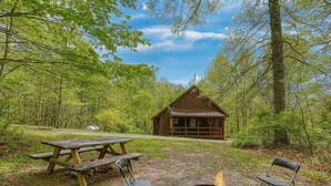 Een chalet omgeven door bomen, met een picknicktafel, stoelen en een vuurkuil.