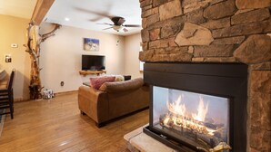 Una sala de estar con chimenea de piedra, ventilador de techo, televisor y piso de madera.