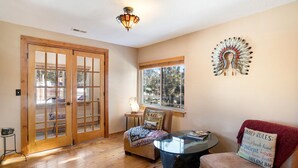 Una sala de estar con piso de madera, una mesa de centro de vidrio, un sofá con cojines, una silla, un ventanal y un elemento decorativo en la pared.