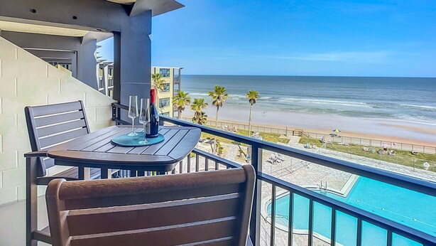 A balcony with a table and chairs, overlooking a beach and a swimming pool.