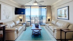 A spacious living room with a large window offering a view of the ocean, a ceiling fan, and a flat-screen TV mounted on the wall.