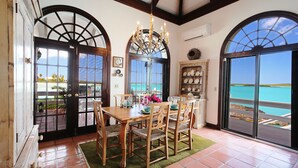 Un comedor con una mesa de madera, sillas y una araña. Se ven amplias ventanas con vista al océano.