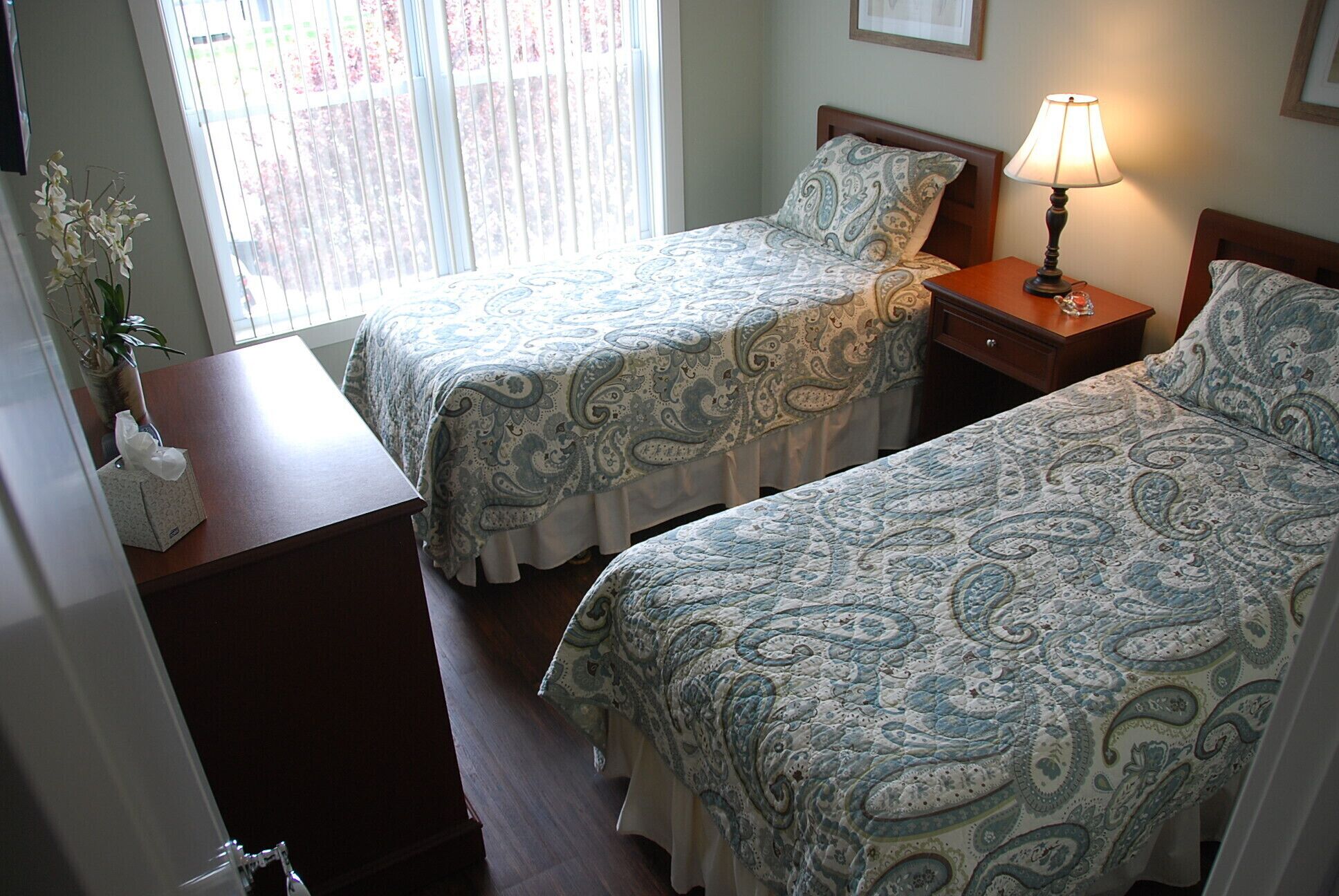 A room with two beds, each with a patterned bedspread, a bedside table with a lamp, and a window with blinds.