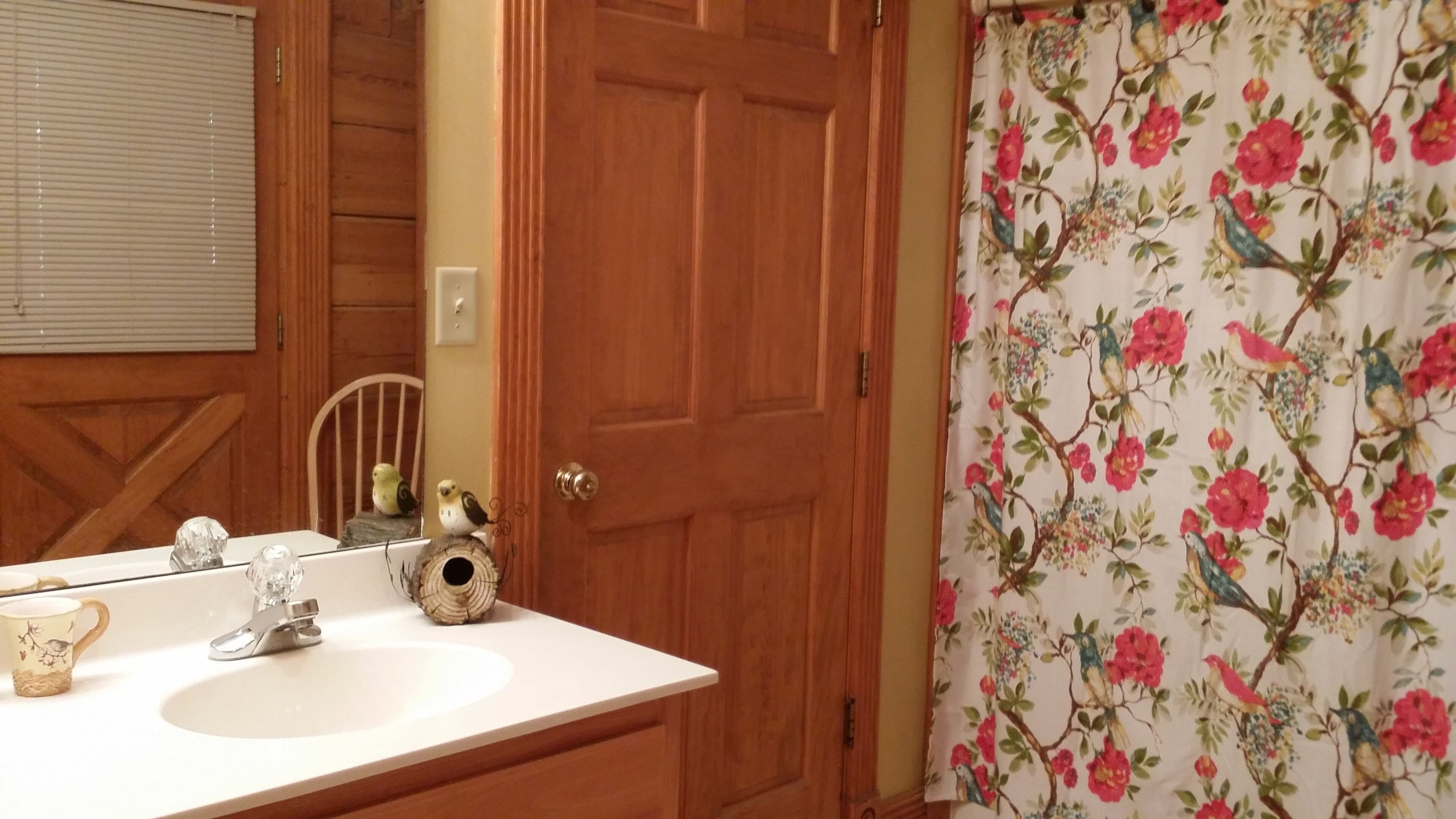 Un baño con puerta de madera, un lavamanos blanco, un espejo y una cortina de ducha con estampado floral.