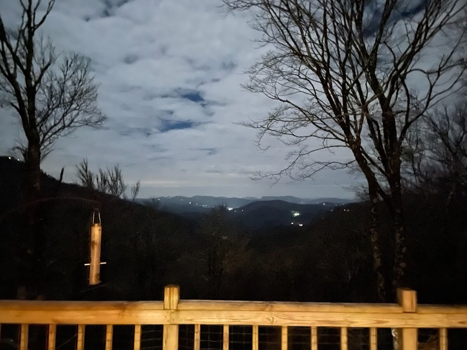 Une vue nocturne depuis une terrasse en bois donnant sur une vallée, avec des lumières scintillantes au loin, des arbres sans feuilles et un ciel nuageux.