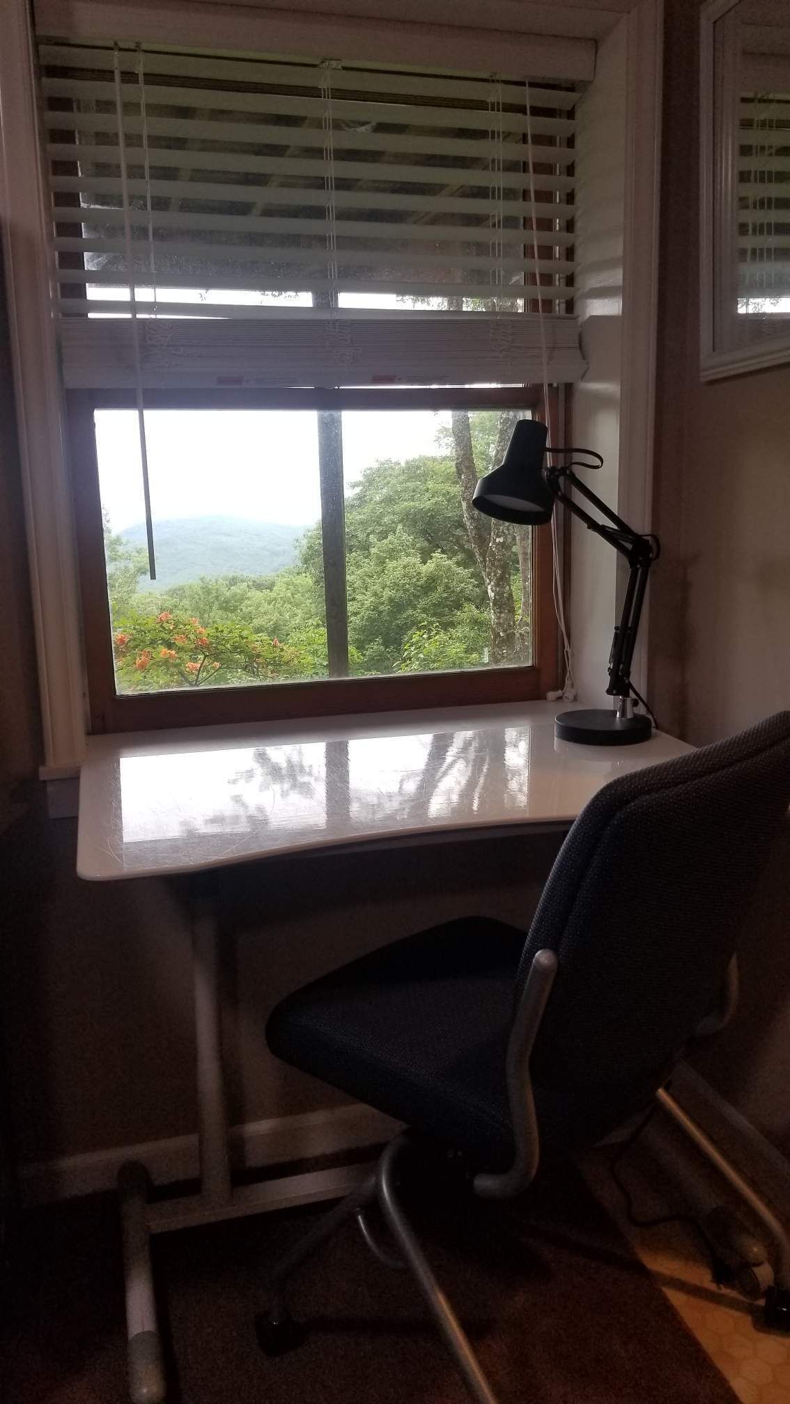 A desk with a computer monitor, a lamp, and a chair in front of a window with blinds.