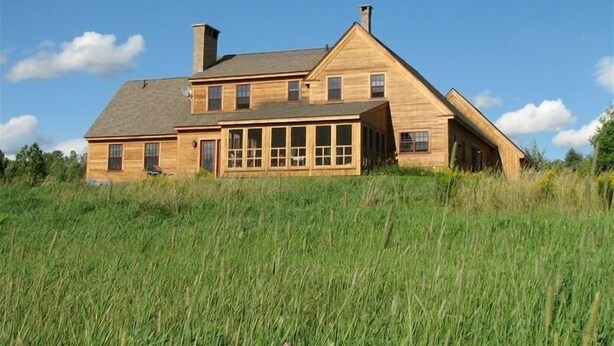 Een houten huis met twee verdiepingen, een puntige dakkapel en schoorsteen, omringd door hoog gras.