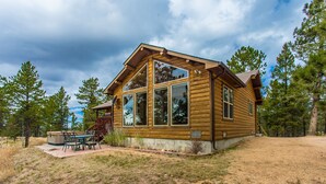 Una cabaña de troncos con ventanas grandes, un patio con mobiliario exterior y vistas al bosque circundante.