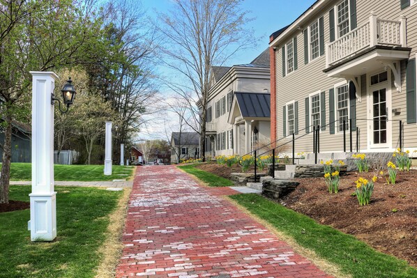 A brick pathway leading to a row of townhouses with green shutters and a well-maintained lawn.