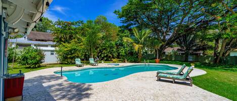 Una piscina con sillones de descanso, rodeada de exuberante vegetación y un patio de piedra.