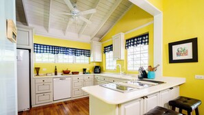 Una cocina con armarios blancos, una isla central con taburetes, un refrigerador y una ventana con un toldillo a cuadros.