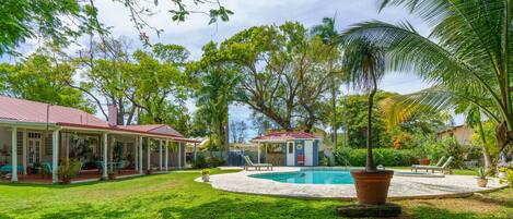Un'area piscina con zona relax coperta, un piccolo edificio e vegetazione lussureggiante.