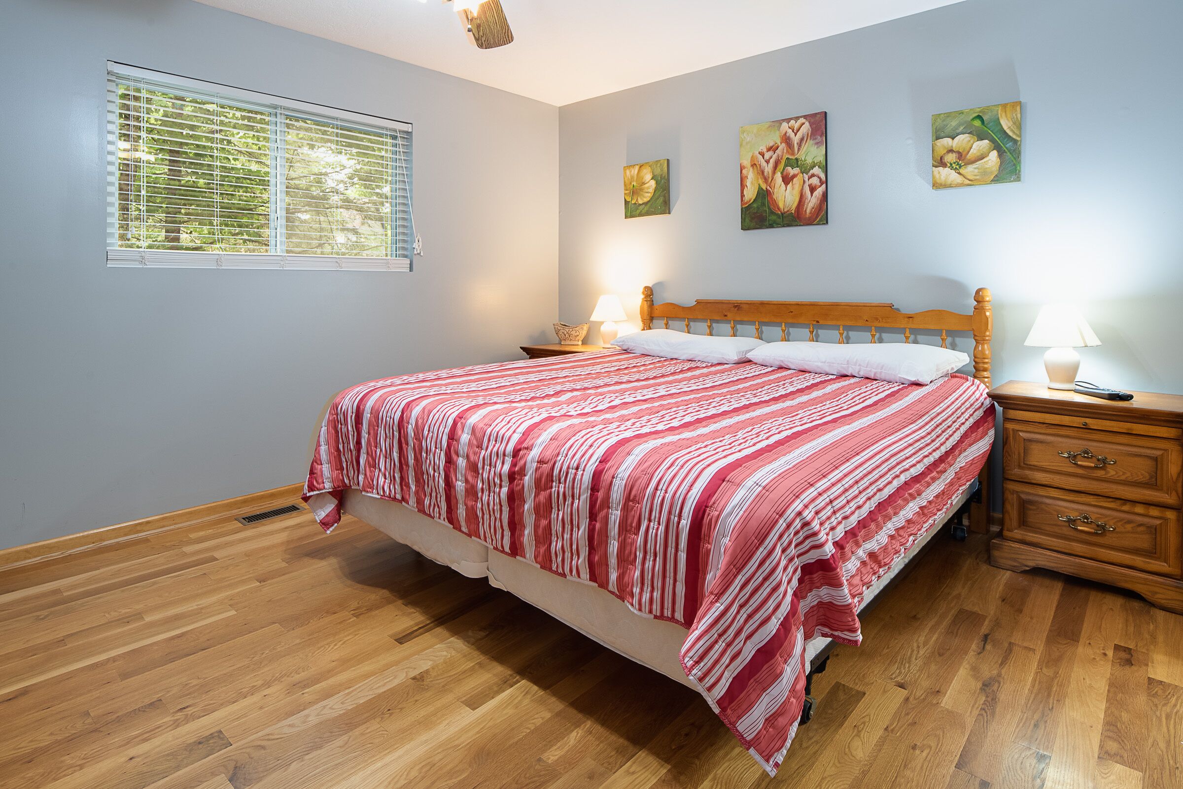 A bedroom with a wooden bed, two bedside tables, a window with blinds, and floral wall art.