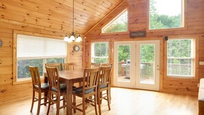 A wooden cabin interior with a dining table, chairs, a clock, and a 'WELCOME' sign.