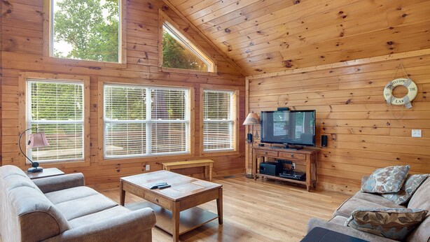 A cozy cabin interior with a wooden ceiling, large windows, a television, and a sofa.