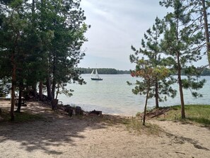 A sandy beach with trees, a sailboat on the water, and a distant shoreline.