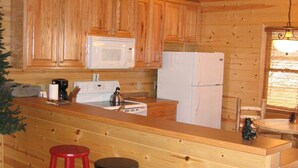 A cabin-style kitchen with wooden cabinets, a white refrigerator, a microwave, and a stove. There are two stools and a couch with pillows.