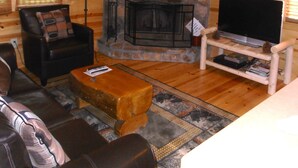 A cozy log cabin interior with a stone fireplace, a comfortable leather sofa, a television on a wooden stand, and a ceiling fan.