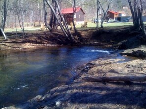 Un ruisseau aux eaux claires serpente à travers une zone boisée, avec un bâtiment à toit rouge visible au loin.