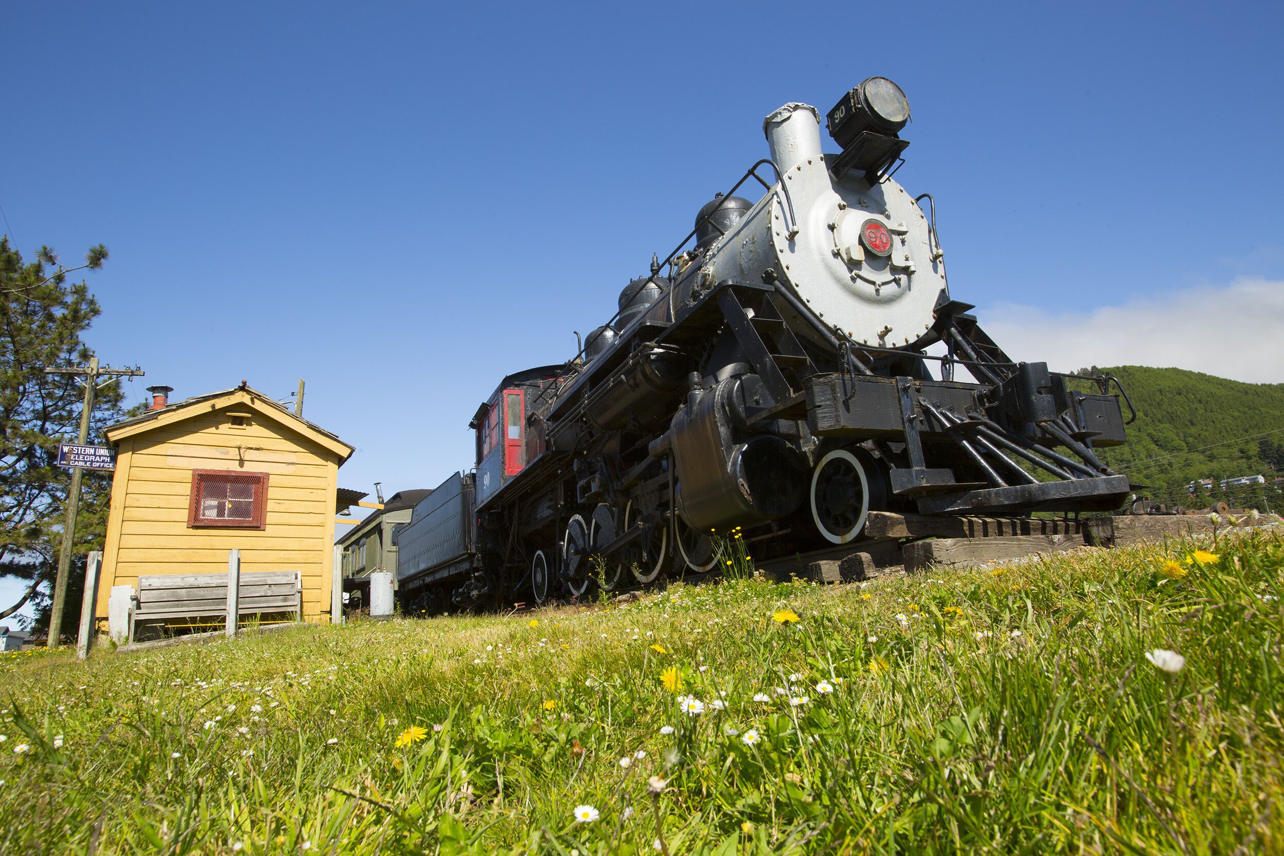 Une locomotive à vapeur sur une colline herbeuse, avec un petit bâtiment jaune à proximité.